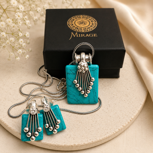 A turquoise and silver jewelry set by Mirage, including a rectangular pendant necklace, matching drop earrings, and a black branded gift box, arranged on a beige surface with decorative fabric and small white flowers.
