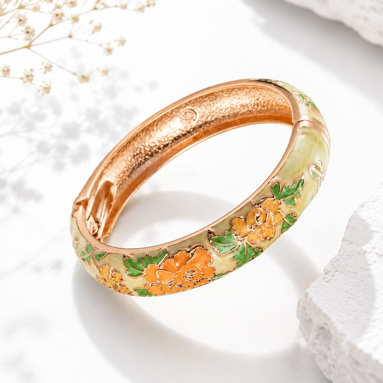 A rose gold bangle bracelet with hand-painted orange flowers and green leaves, displayed on a textured white surface with soft shadows.