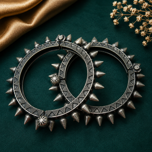 Two ornate silver-toned spiked hoop earrings arranged overlapping on a dark green textured surface with a gold fabric corner and small dried flowers in the upper right.