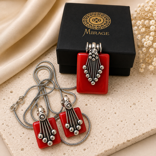 A red rectangular pendant necklace and matching rectangular stud earrings with silver-toned beaded fringe, displayed with a black Mirage jewelry box on a beige stone surface with cream fabric and dried flowers.