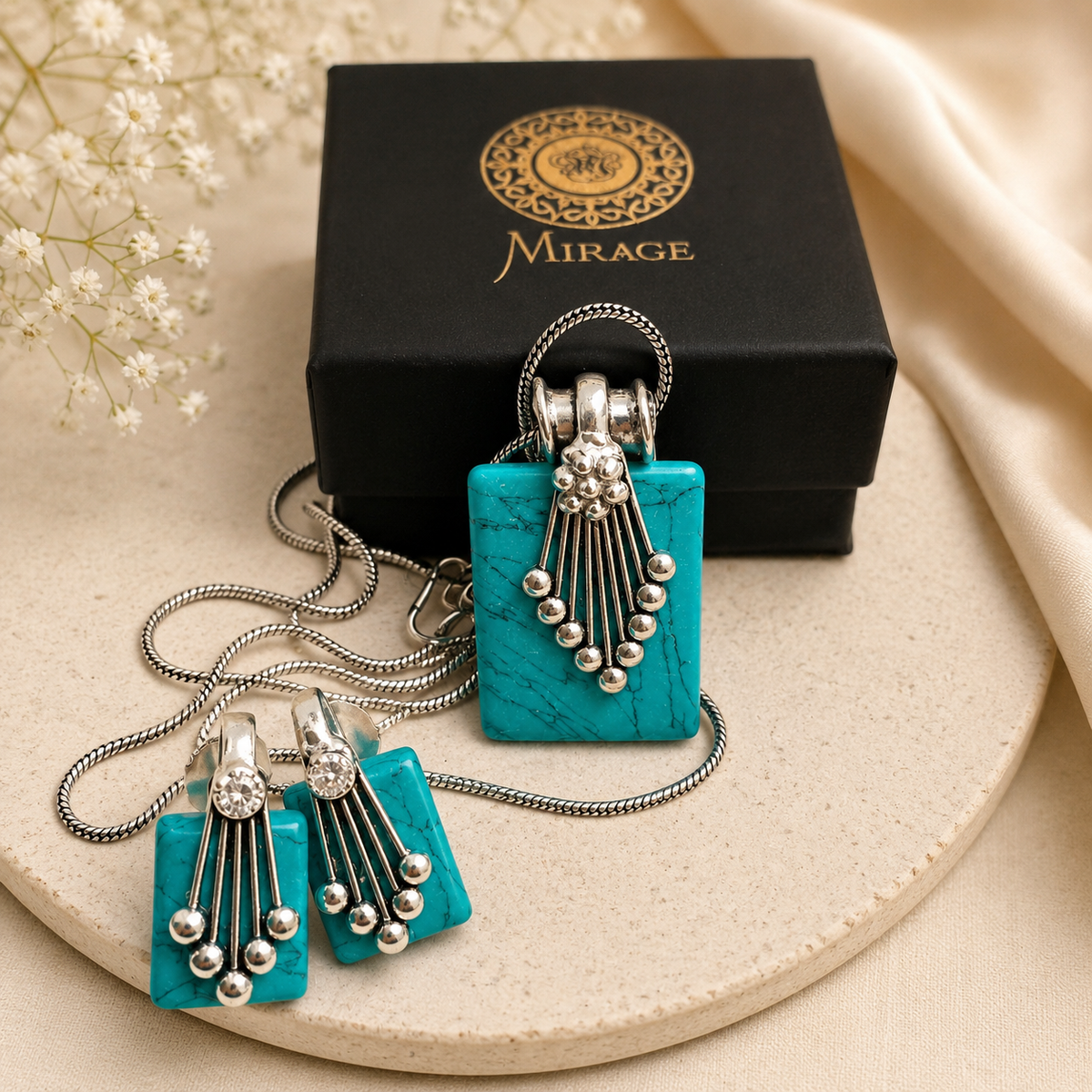 A turquoise and silver jewelry set by Mirage, including a rectangular pendant necklace, matching drop earrings, and a black branded gift box, arranged on a beige surface with decorative fabric and small white flowers.